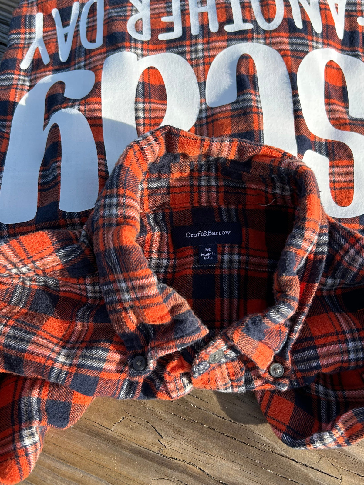 A folded orange and navy plaid flannel shirt with large white text reading 'stay another day' laid on a weathered wooden surface.